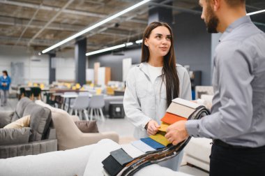 Mobilya galerisinde erkek bir satış danışmanıyla kumaş seçeneklerini tartışan kadın müşteri, yeni ev alımları için malzeme seçiyor.