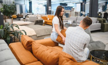 Genç çift parlak bir mobilya galerisinde kanepeyi test ediyor, birlikte oturuyor ve yeni modern daireleri için parçalar seçiyorlar.