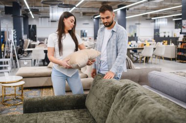 Genç çift alışveriş yapıyor, kanepe dokusunu ve yastık tasarımını büyük modern mobilya galerisinde karşılaştırıyor.