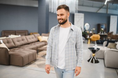 Genç adam modern bir mobilya mağazasında duruyor, iç tasarım ve ev dekorasyonu için yeni bir kanepe seçiyor.