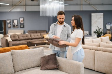 Young couple shopping for a sofa in a modern furniture store, selecting a pillow for their living room design