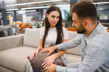 Young couple sitting on a sofa, feeling and discussing a decorative pillow in a modern furniture showroom