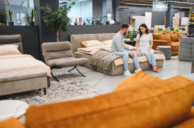 Young couple sitting on a bed, discussing new furniture for their home, making design choices in a large retail store