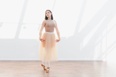 Genç balerin bir dans stüdyosunda bale yapıyor bej bir tayt ve bale giyip, duruşunu ve zarafetini gösteriyor.