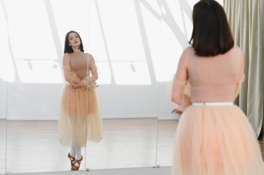 Genç bayan balerin parlak bir dans stüdyosunda poz vererek kendini düzeltmek ve geliştirmek için ayna kullanıyor.