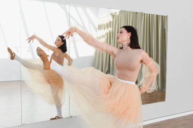 Genç bayan balerin dans stüdyosunda yüksek tekme pratiği yapıyor. Aynanın karşısında zarif bir etek giyiyor.