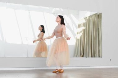 Genç bayan balerin dans stüdyosunda dans figürleri yapıyor. Aynanın önünde etek giyiyor.