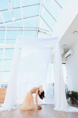 Genç bayan balerin, parlak bir dans stüdyosunda büyük pencerelerden süzülen ışıkla zarif bir geri dönüş pratiği yapıyor.