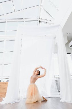 Genç balerin beyaz perdelerle güneşli bir stüdyoda diz çöküp zarif bir dans gösterisi yapıyor.