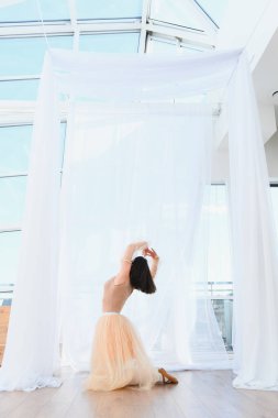 Şeftali rengi etekli genç bir kadın beyaz perdeli güneşli bir stüdyoda zarif bir bale dansı yapıyor.