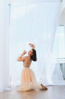 Genç zarif balerin beyaz perdeleri ve ahşap zemini olan parlak bir stüdyoda kollarıyla diz çöküp dans ediyor.