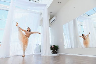 Genç bayan balerin, parlak bir dans stüdyosunda bacak uzatma alıştırması yapıyor. Zarif duruşunu yansıtan bir aynayla.