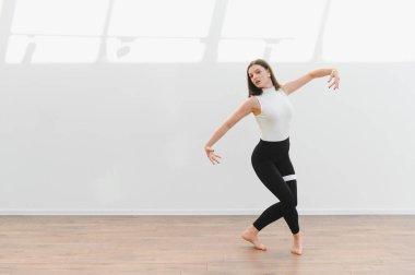 Dans öğrencisi, ahşap zemini ve beyaz duvarları olan parlak bir stüdyoda modern bir dans koreografisi uyguluyor.