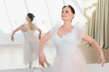 Beyaz etekli yaşlı balerin dans stüdyosunda bale hareketleri yapıyor. Yansıması hareketlerini yansıtıyor.