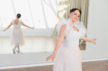 Beyaz etekli yaşlı balerin bir bale stüdyosunda zarif bir şekilde dans ediyor. Yansıması hareketlerini yansıtıyor.