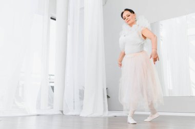 Yaşlı balerin zarif bir şekilde dans ve aktif yaşlanmanın güzelliğini gösteren beyaz bir stüdyoda bale hareketleri sergiliyor.