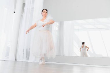 Kıdemli balerin, parlak beyaz bir stüdyoda bale hareketleri yapıyor, yaşlılıkta dans için zarafet ve tutku sergiliyor.