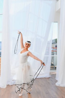 Yaşlı balerin beyaz etek ve sivri ayakkabılar giyerek parlak bir stüdyoda kurdelelerle dans ediyor.