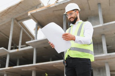 Gülümseyen mühendis güvenlik donanımı giyiyor, inşaat planlarını gözden geçiriyor, proje geliştirme çalışmalarını denetliyor.
