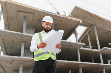 Erkek mimar yeni inşaat alanının planlarını inceliyor. Kasket ve güvenlik yeleği giyiyor. Gelişmeyi denetliyor.