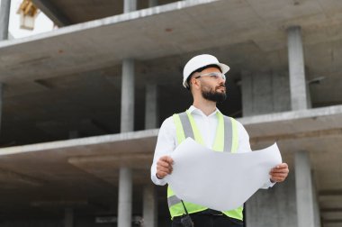 Güvenlik yeleği ve şapka takan genç erkek mühendis inşaat alanının planlarını dikkatle inceliyor.