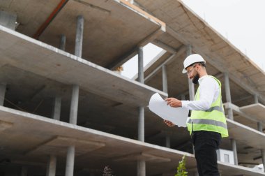Sert şapka ve güvenlik yeleği giyen genç erkek mühendis beton inşaat alanındaki bina planlarını inceliyor.