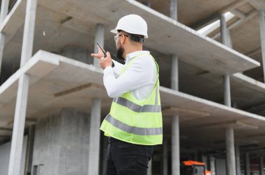 Erkek mühendis çelik yelek giyiyor, inşaat alanında telsizle iletişim kuruyor. Geliştirmeyi denetle