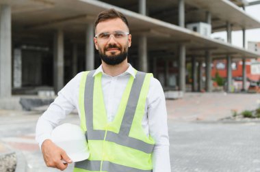 Güvenlik yeleği ve temiz gözlük takan profesyonel bir adam, elinde bir şapka, geleceğin bina yapısının önünde duruyor.