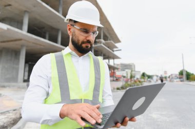 Mühendis ya da mimar sıkı bir şapka ve güvenlik yeleği giyiyor. İnşaat alanında dizüstü bilgisayarla çalışıyor.