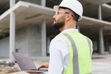 Güvenlik yeleği ve başlık takan erkek mühendis inşaat ve inşaat alanındaki dizüstü bilgisayarlara bakıyor.