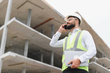 Mühendis çalışıyor, akıllı telefondan konuşuyor, inşaat alanında dizüstü bilgisayar tutuyor. İnşaatta güvenlik ve teknoloji