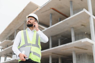 Sert şapka ve güvenlik yeleği giyen, dizüstü bilgisayarı olan bir adam inşaat alanında ilerleme kaydetmeyi tartışıyor.