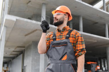 Erkek inşaat işçisi, başlık ve güvenlik gözlüğü takıyor. İnşaat alanında telsizle iletişim kuruyor.