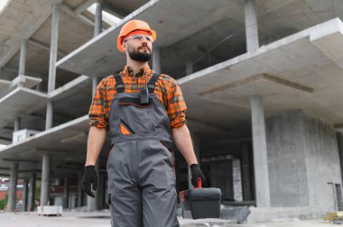 Erkek inşaat işçisi, kasket ve güvenlik gözlüğü takıyor, ticari bir inşaat alanında alet çantası tutuyor.