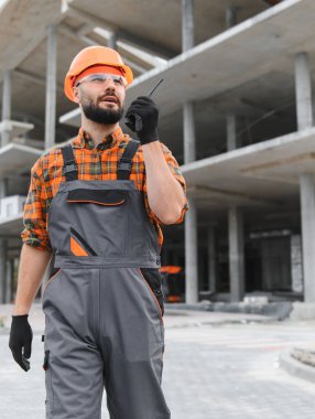 İnşaat işçisi, inşaat başlığı ve güvenlik gözlüğü takıyor. Bina inşaatında iletişim kurmak için telsiz kullanıyor.
