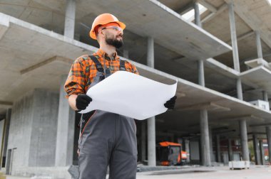 İnşaat işçisi inşaat planlarına bakıyor, büyük beton inşaat alanındaki inşaat çalışmalarını denetliyor.