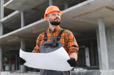 İnşaat işçisi, başlık ve güvenlik gözlüğü takıyor, elinde bir plan tutuyor, inşaat alanında ilerleme kaydediyor.