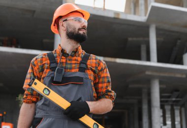 Kasketli ve tulumlu işçiler ruh seviyesinde modern bir şantiyeye bakıyor.