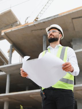 Titiz mühendis elinde bina planı tutuyor, inşaat projesini planlıyor, çelik yelek ve şapka takıyor.