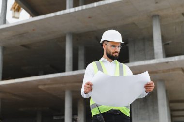 Erkek mühendis ya da mimar inşaat alanındaki bina planlarını inceliyor, çelik yelek ve şapka giyiyor.