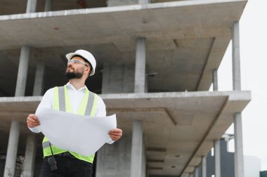 Sıkı bir şapka ve güvenlik yeleği giyen genç bir mühendis elinde planlar, beton bir inşaat alanına bakan...