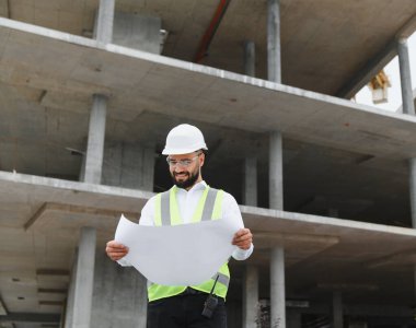 Mühendis gülümsüyor, aktif bir inşaat alanındaki bina planlarını inceliyor, güvenlik başlığı ve yelek giyiyor.