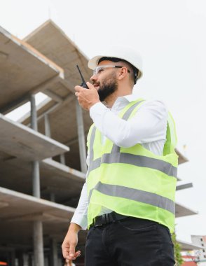 İnşaat mühendisi çelik yelek ve miğfer takıyor. İnşaat alanında telsiz kullanıyor.