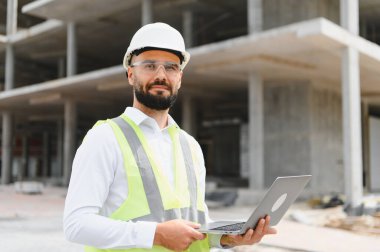 Başlık ve güvenlik yeleği giyen mühendis dizüstü bilgisayar tutuyor ve inşaat alanındaki gelişmeleri inceliyor.