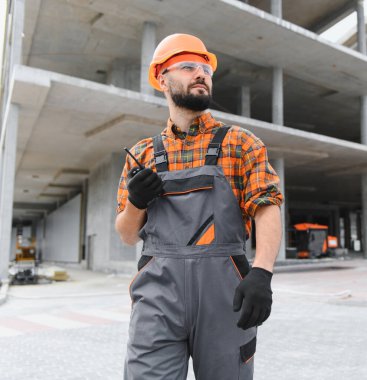 Mühendis ya da ustabaşı, sağlam şapka ve güvenlik gözlüğü takıyor, beton bir inşaat arka planında bir telsiz tutuyor.