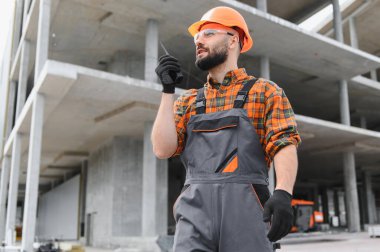 İnşaat işçisi telsizle iletişim kuruyor, inşaat alanında şapka ve güvenlik gözlüğü takıyor.