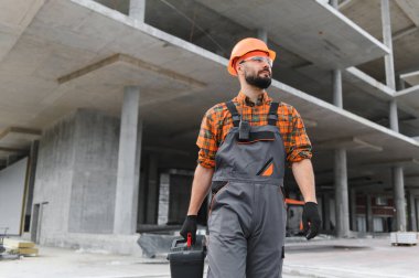 Koruyucu üniforma, şapka ve güvenlik gözlüğü takan, beton inşaat alanında alet çantası taşıyan bir erkek işçi.