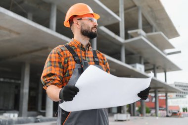 İnşaat alanındaki inşaat planlarını inceleyen kasklı ve güvenlik gözlüklü genç mühendis ya da mimar.