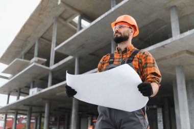 İnşaat mühendisi, başlık ve güvenlik gözlüğü takıyor, elinde bir plan tutuyor, planlama yaparken ileriye bakıyor.