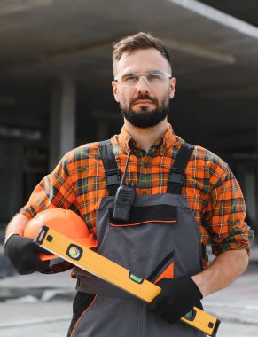 Kaliteli inşaat işçisi inşaat alanında çalışıyor, kafasında şapka ve ruh seviyesi var, koruyucu giysiler giyiyor.